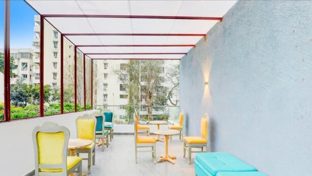 A covered outdoor seating area with chairs and tables, a blue bench, and a view of buildings and trees. - The Grand Homes (Lucknow)