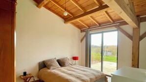 A bedroom with a wooden ceiling, a bed with a beige blanket, a red lamp, and a view of the outdoors through a large window.