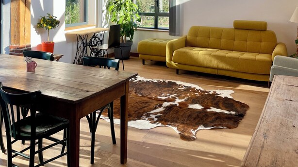 A spacious loft with a high wooden ceiling, large windows, a yellow sofa, a dining table, and a cowhide rug.