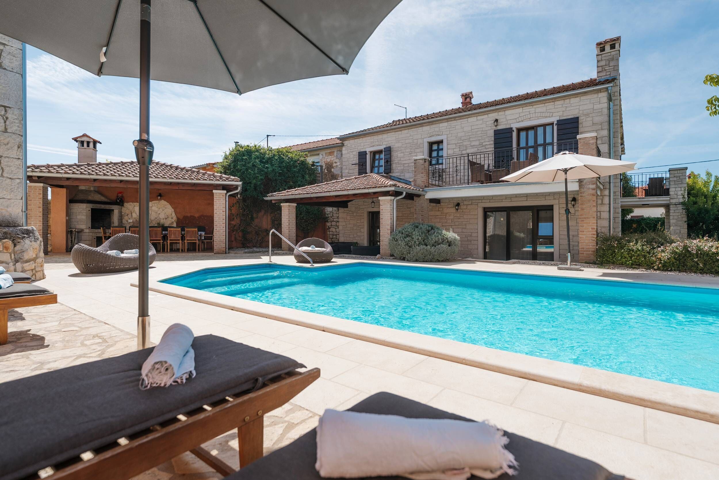 Une piscine avec des chaises longues et des parasols, un bâtiment en pierre avec un toit en tuiles et une cheminée en pierre.