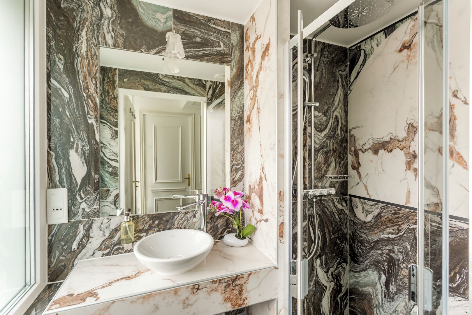 A bathroom with a marble vanity, a white sink, a pink orchid, and a shower with marble walls.