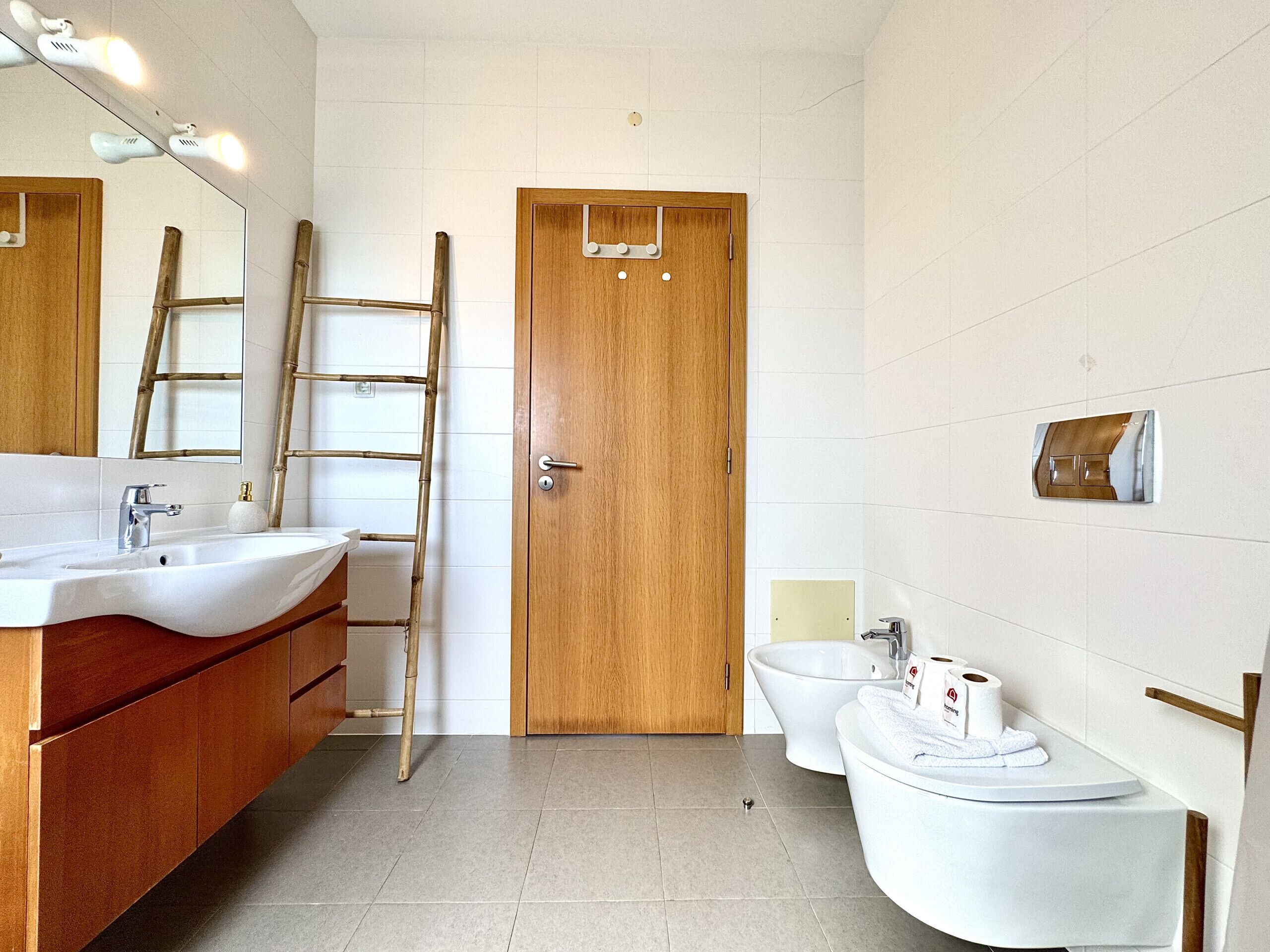 A bathroom with a wooden door, a sink, a toilet, and a mirror with lights.