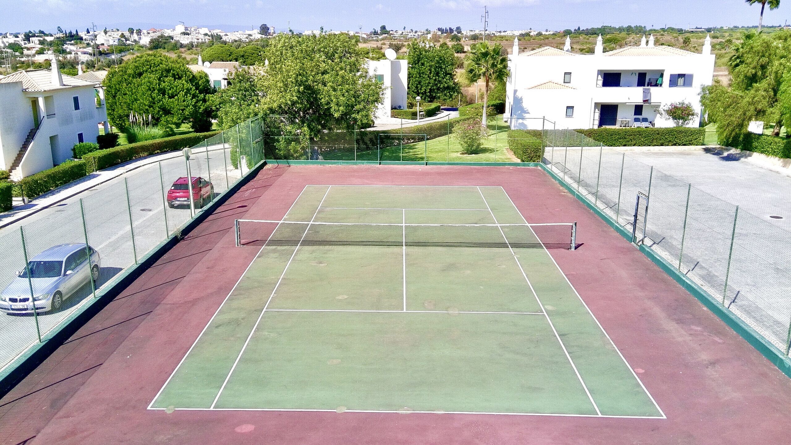 Una cancha de tenis con una red, rodeada por una valla, y junto a una calle con coches estacionados.