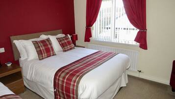A neatly made bed with white linens and a plaid blanket, a wooden nightstand with a lamp, and a window with red curtains.