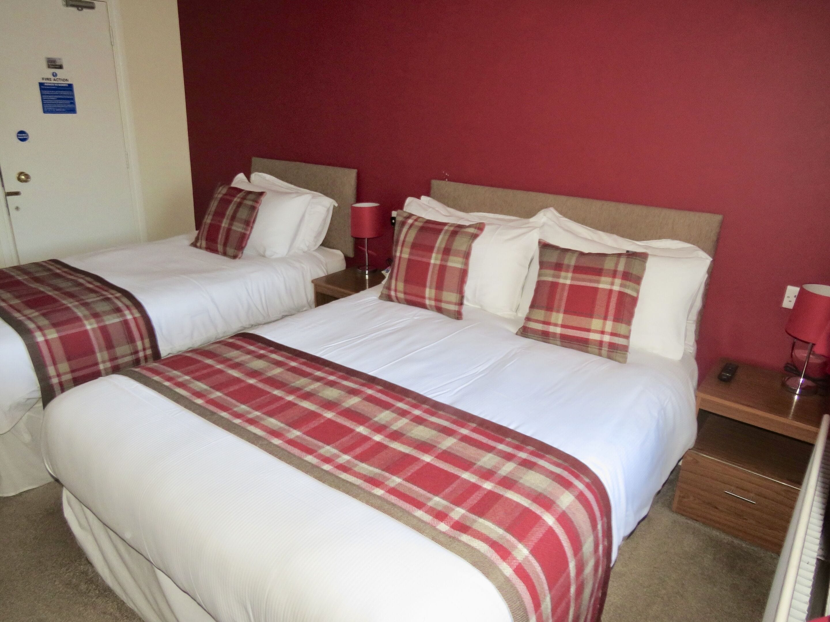 A hotel room with two beds, each with white bedding and red plaid blankets. There is a red wall and a door with a fire action sign.