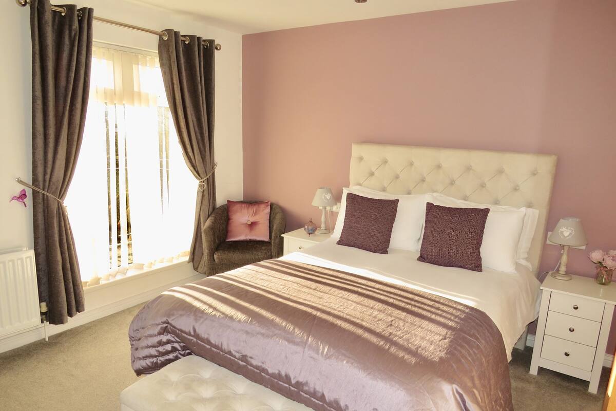 A bedroom with a large bed, a chair, a nightstand, and a window with curtains.
