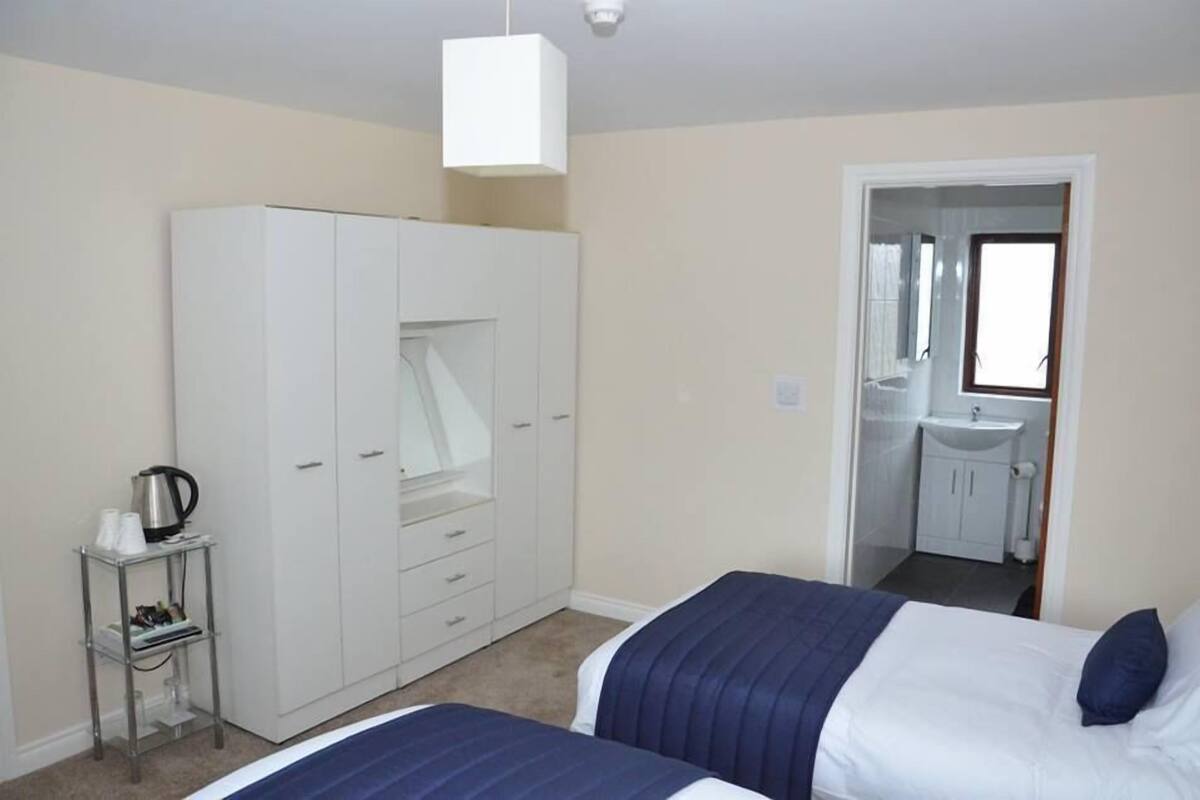 A bedroom with two beds, a wardrobe, a small table with cups and a kettle, and a bathroom visible through an open door.