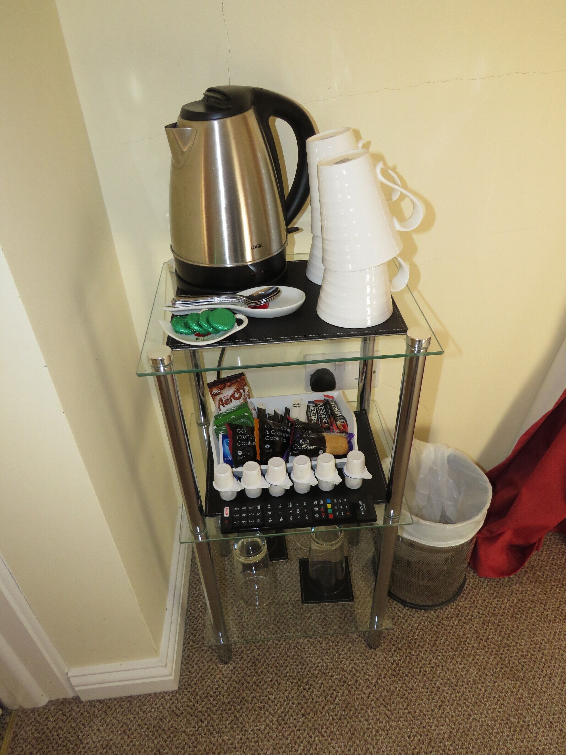 A glass shelf with a kettle, coffee cups, a plate with cookies, a trash bin, and a glass of water.