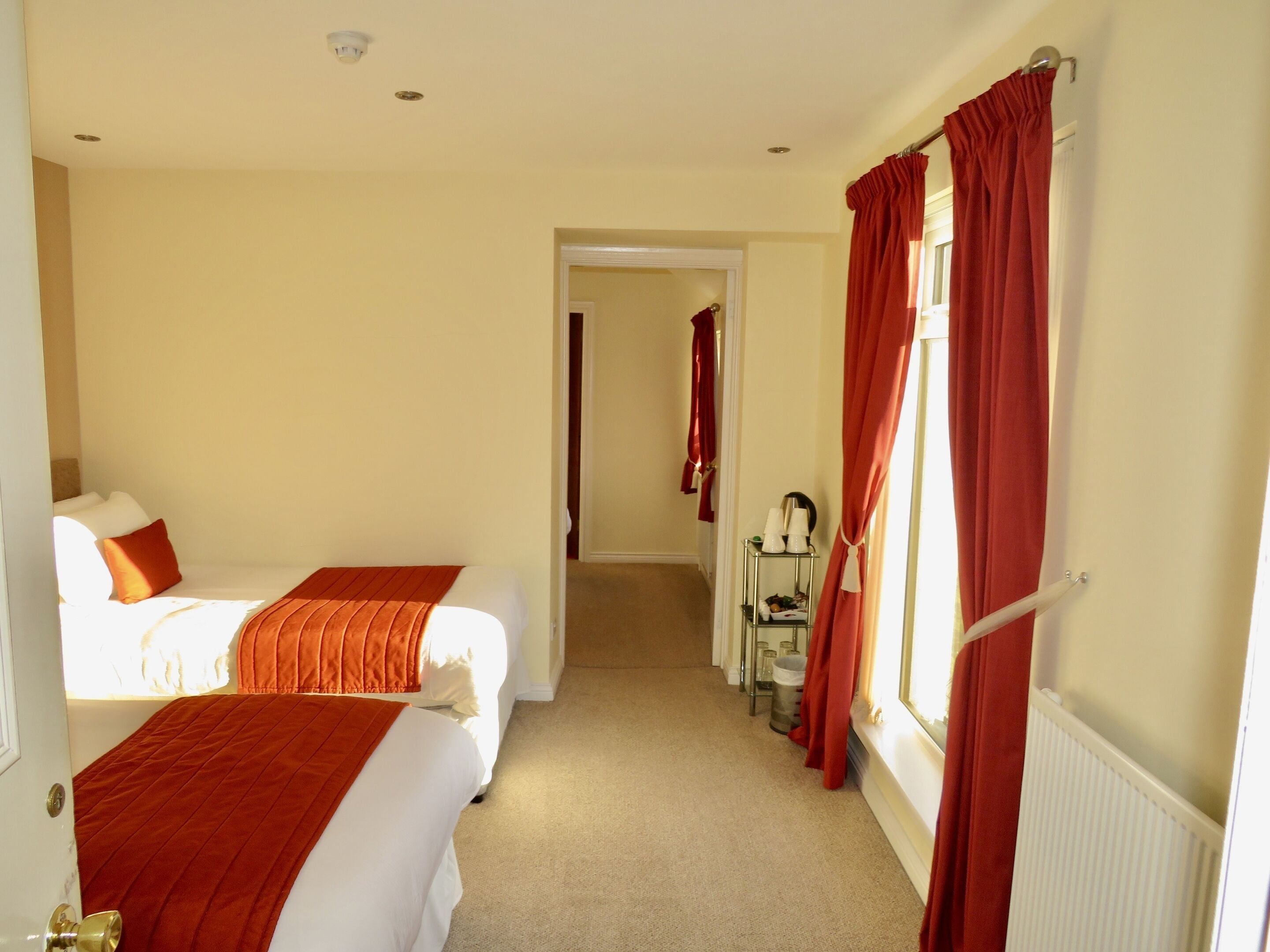 A hotel room with two beds, a hallway, and a window with red curtains.