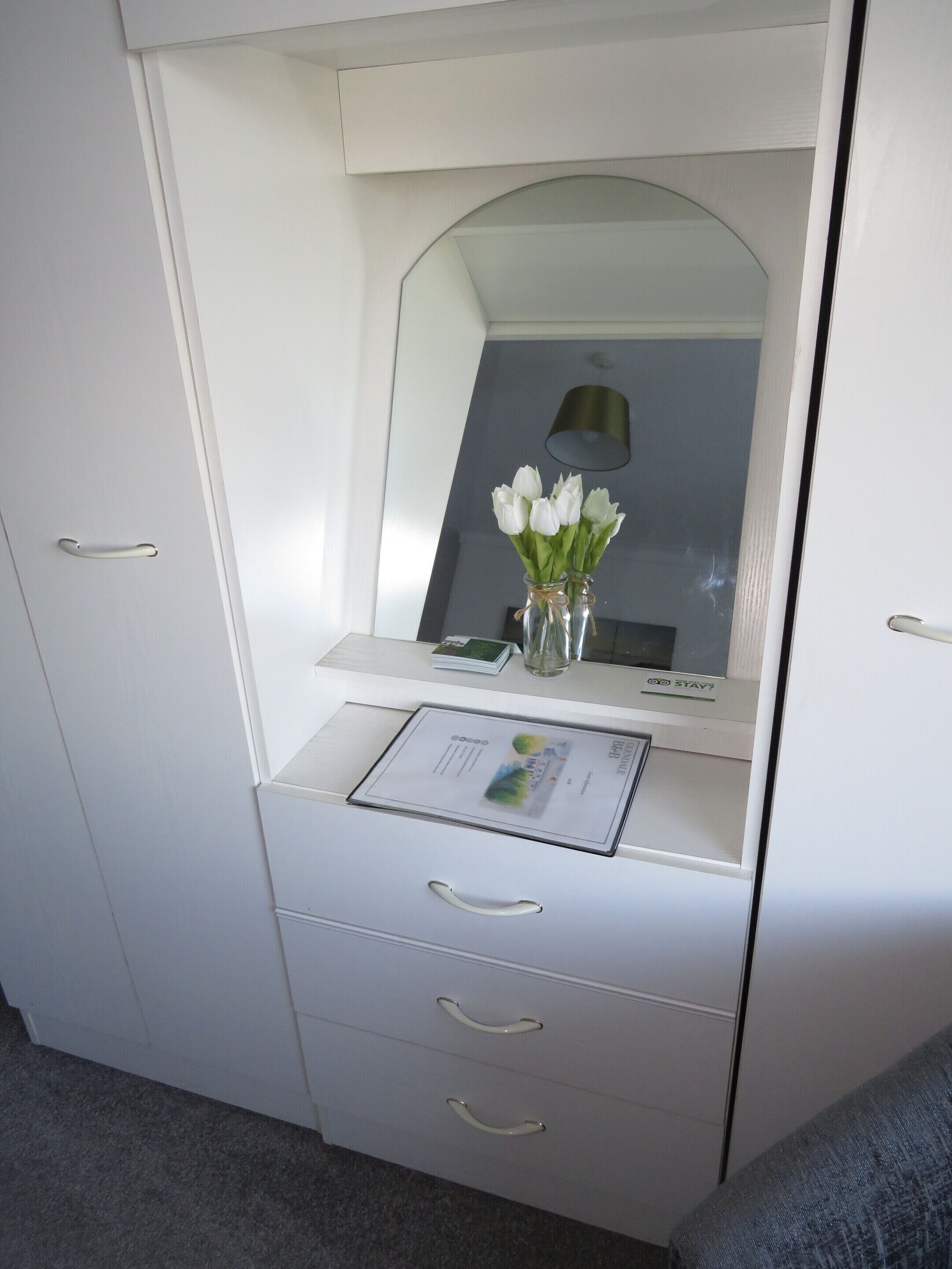 A white wardrobe with a mirror, a small desk with a lamp, and a vase with white flowers.