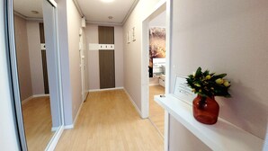 Un couloir avec un plancher en bois, un mur blanc et un miroir. On aperçoit un vase rempli de fleurs sur une étagère.