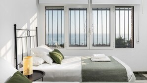A bedroom with a bed, pillows, a bedside table, a lamp, and a view of the sea through large windows.