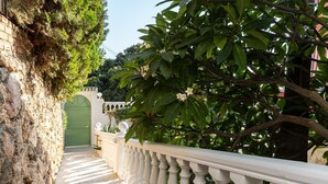 A stone pathway leading to a green gate, surrounded by lush greenery and a white balustrade.