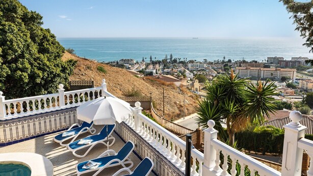 A balcony with sun loungers, a pool, and a view of the sea and town.