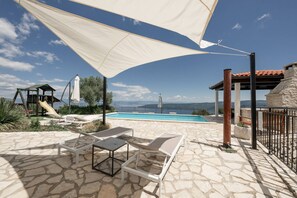 A stone patio with lounge chairs and a pool, overlooking a scenic view with a playground and a stone building. - Villa Country Lady (100311-K1) - Krsan (Krsan)