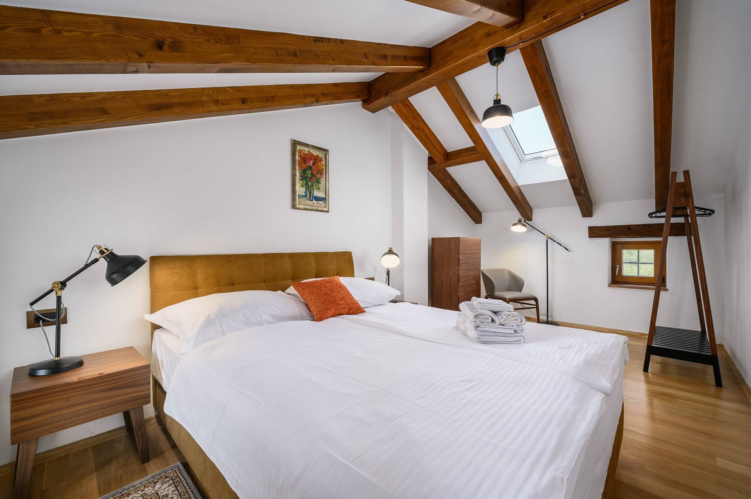 A bedroom with a wooden ceiling, a bed with white bedding, a desk with a lamp, and a painting on the wall.