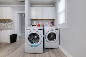 Une buanderie équipée de deux machines à laver à chargement frontal, de meubles blancs, d’un pare-feu en carrelage et d’une poubelle noire.