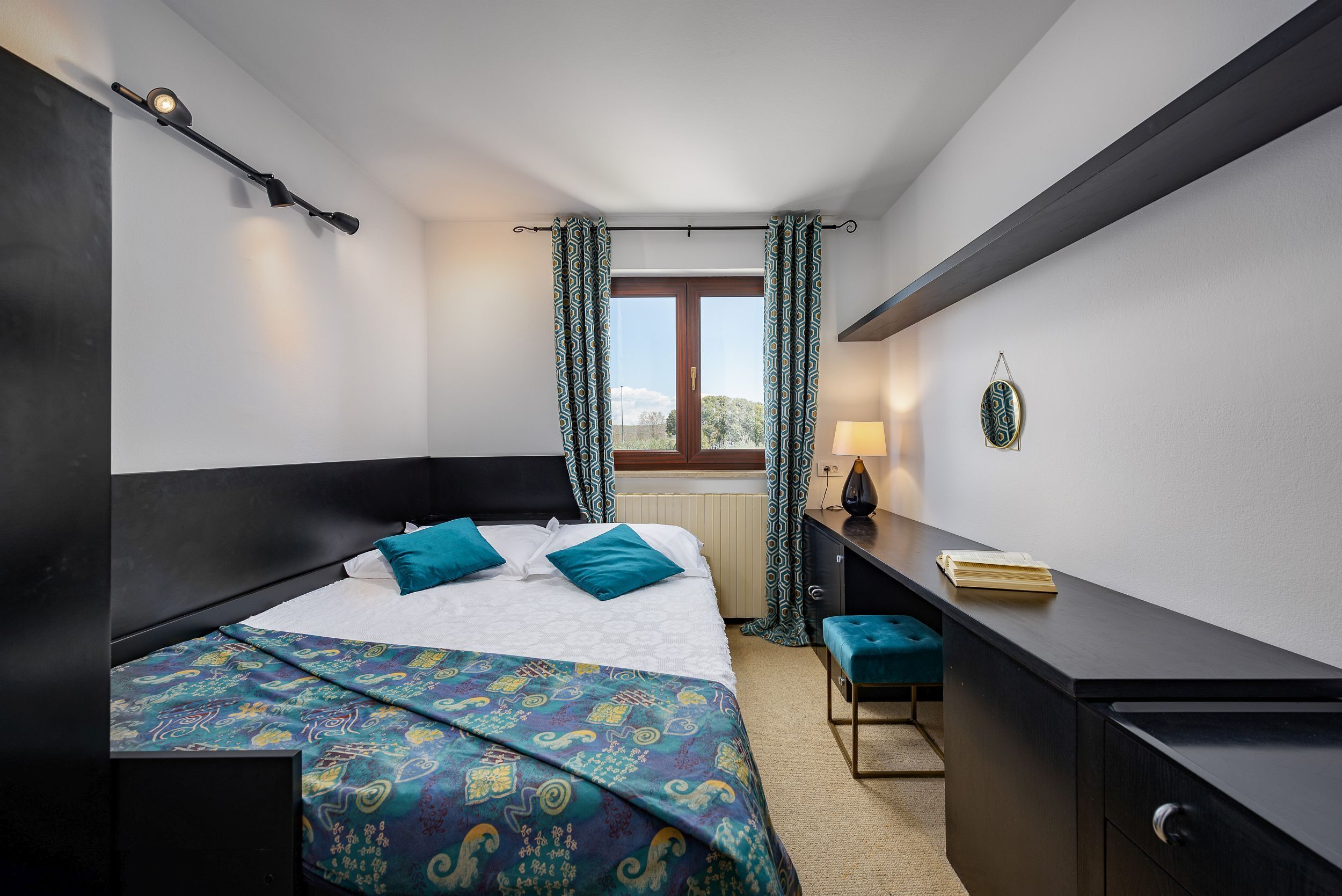 A bedroom with a bed, a desk, a chair, a window with curtains, and a wall-mounted lamp.