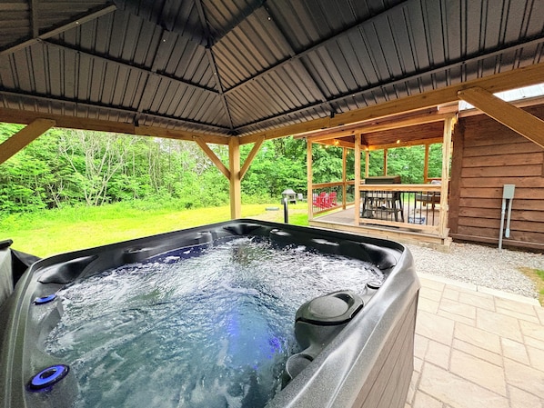 A hot tub with a wooden deck and a covered area, surrounded by greenery.
