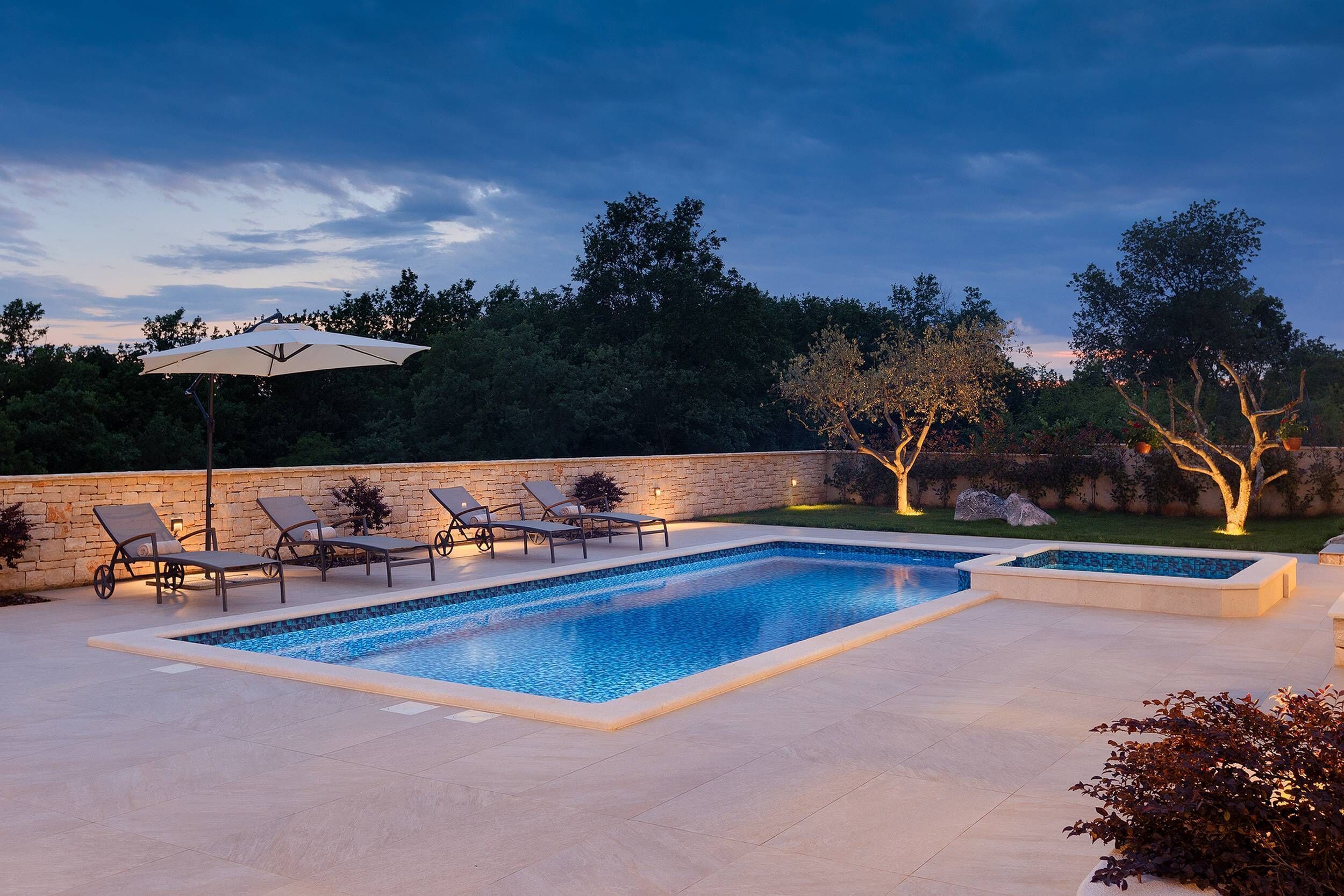 Una piscina rectangular con solárium azulejado, rodeada de sillas de descanso y un gran paraguas.