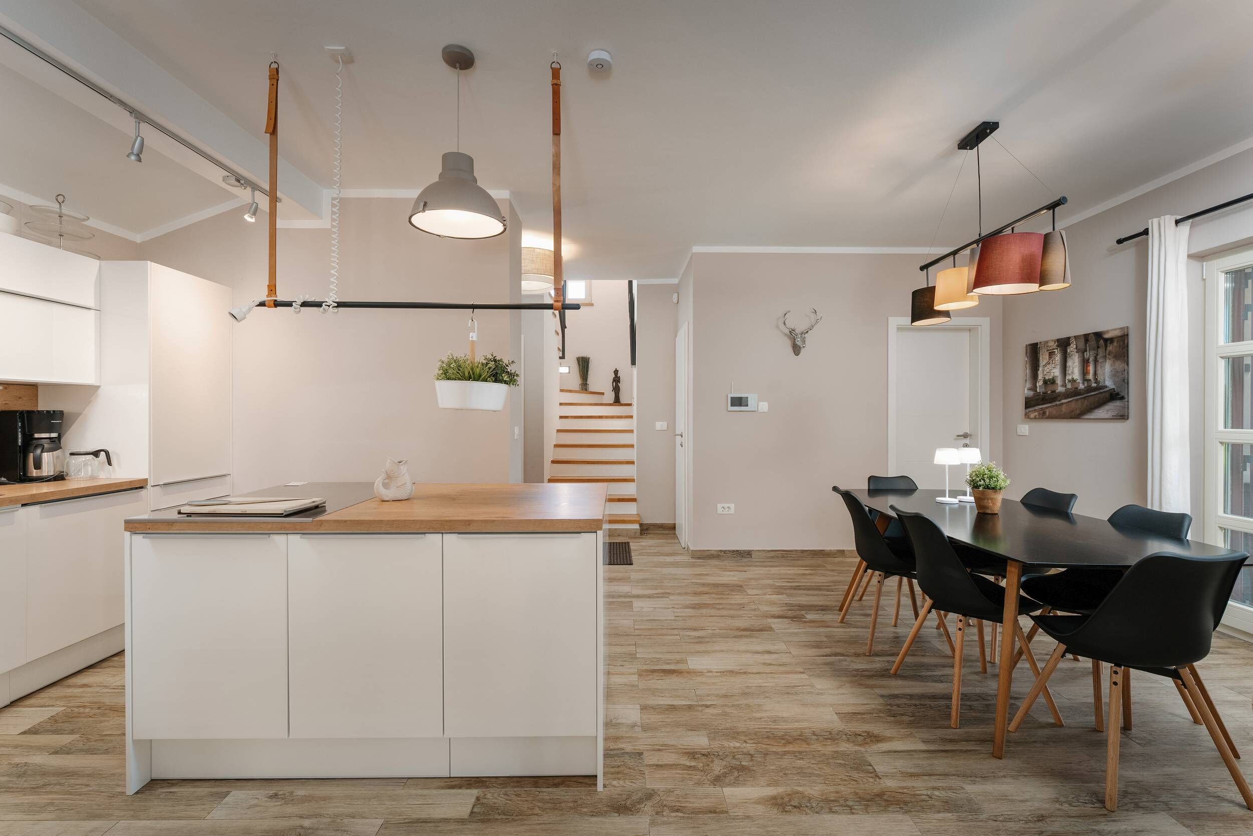 Una cocina moderna con armarios blancos, una isla de madera, luces colgantes y un comedor con sillas negras y una mesa.