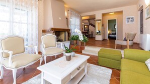A living room with a white sofa, a green sofa, and two white armchairs. There is a coffee table with a plant and decorative plates. The room has a fireplace, a staircase, and a dining area in the background. - Villa Salvea (100391-K1) - Montizana (Montizana)