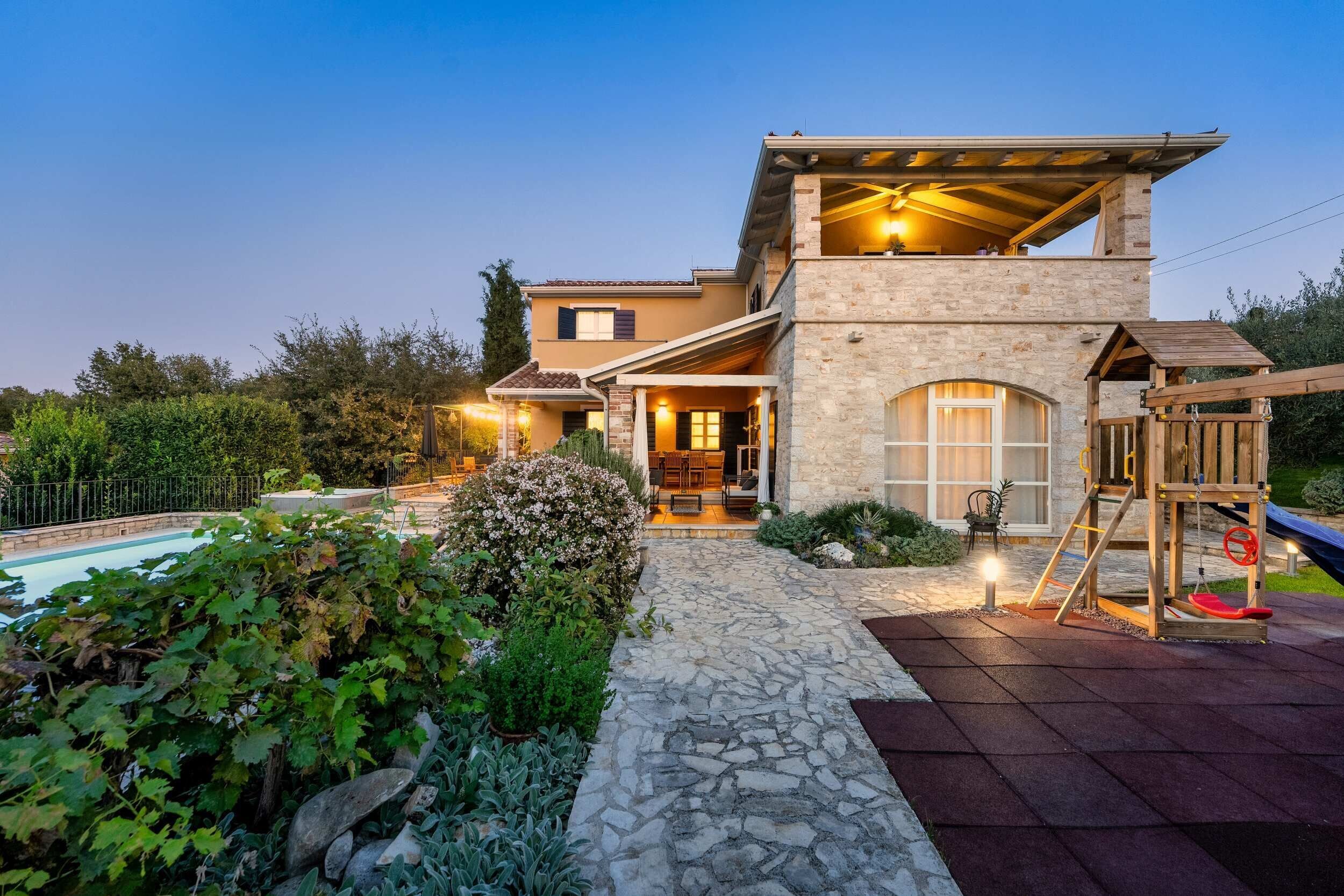 A two-story house with a stone facade, a swimming pool, and a wooden deck with a swing set.