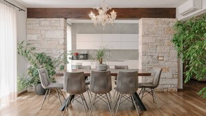 Zona pranzo con tavolo in legno, sedie grigie, un lampadario, pareti in pietra e piante in vaso.