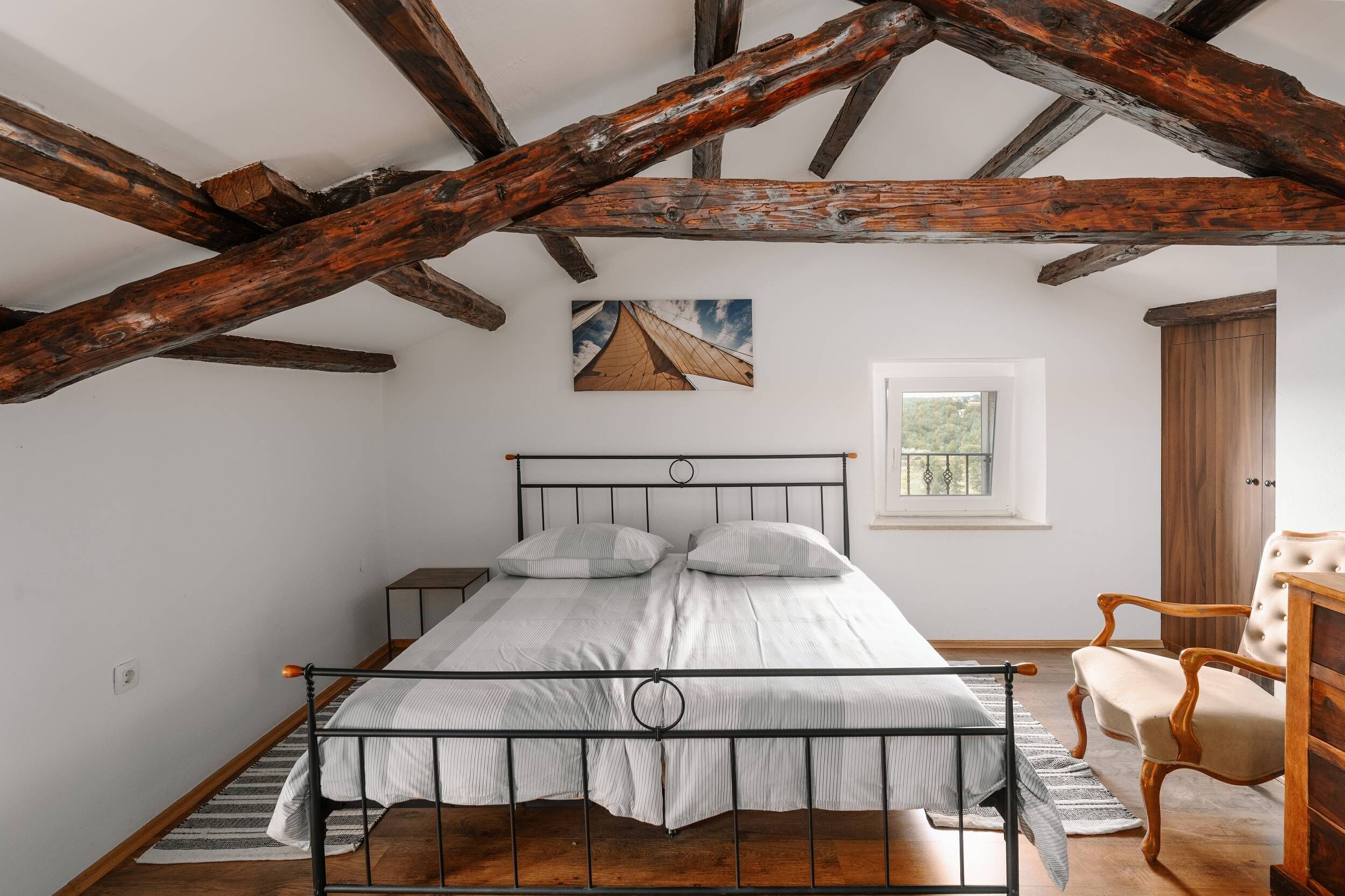 A bedroom with a metal bed frame, a wooden chair, a wooden wardrobe, and a painting on the wall.