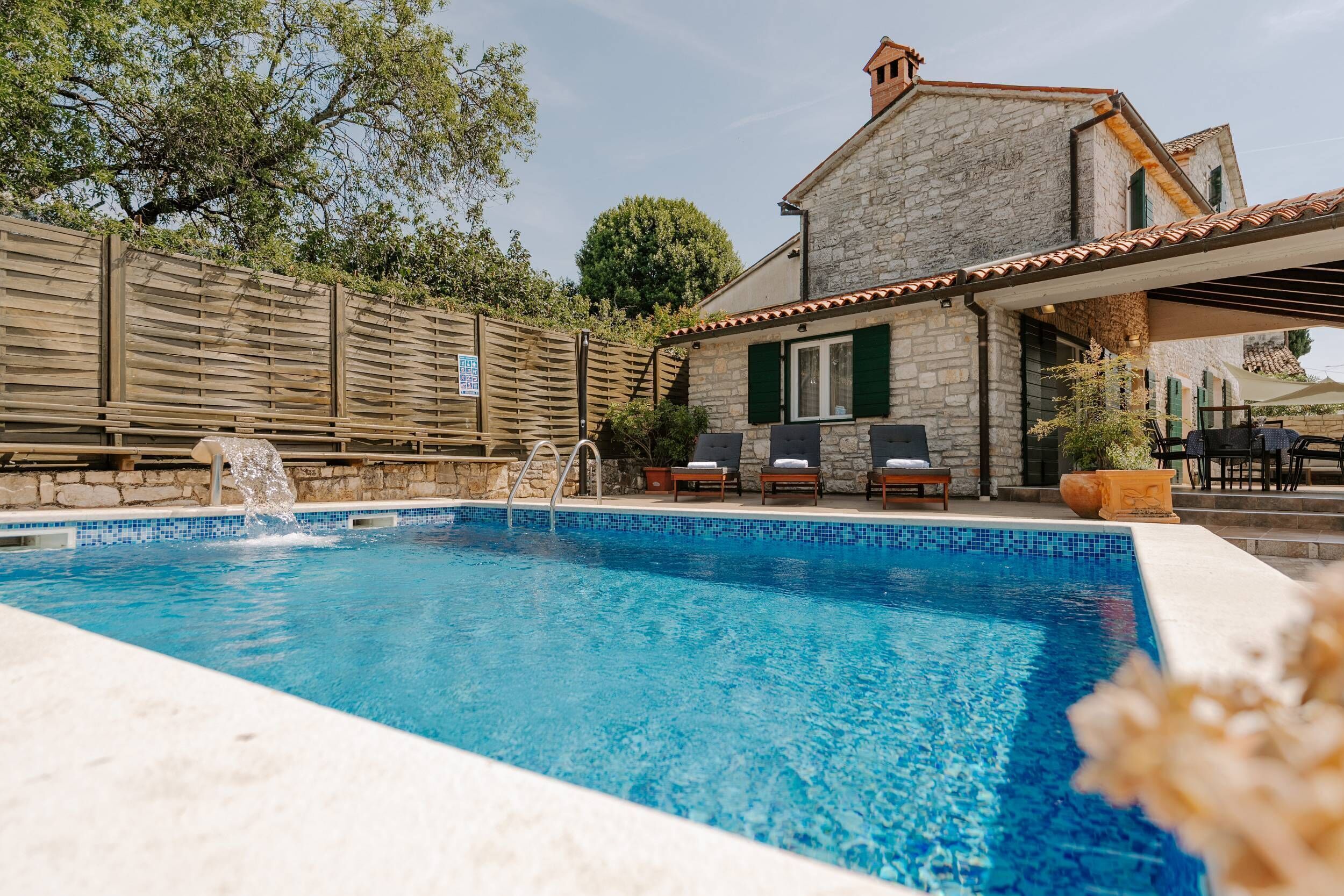 A swimming pool with a stone house in the background, outdoor seating, and a wooden fence.