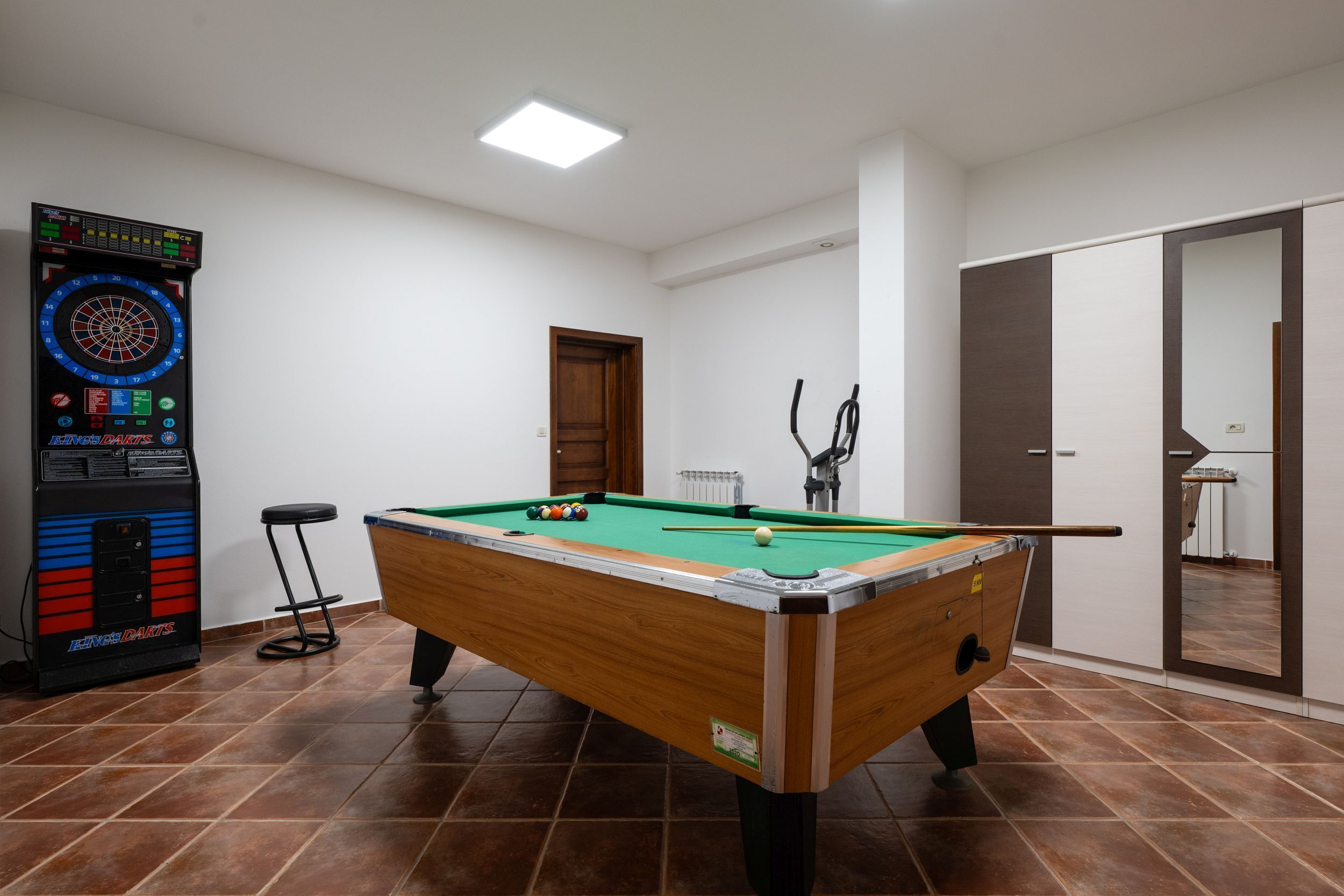 Une table de billard avec des billes et des queues, un jeu de fléchettes et une table de pool dans une pièce dont le sol est carrelé et les murs sont blancs.
