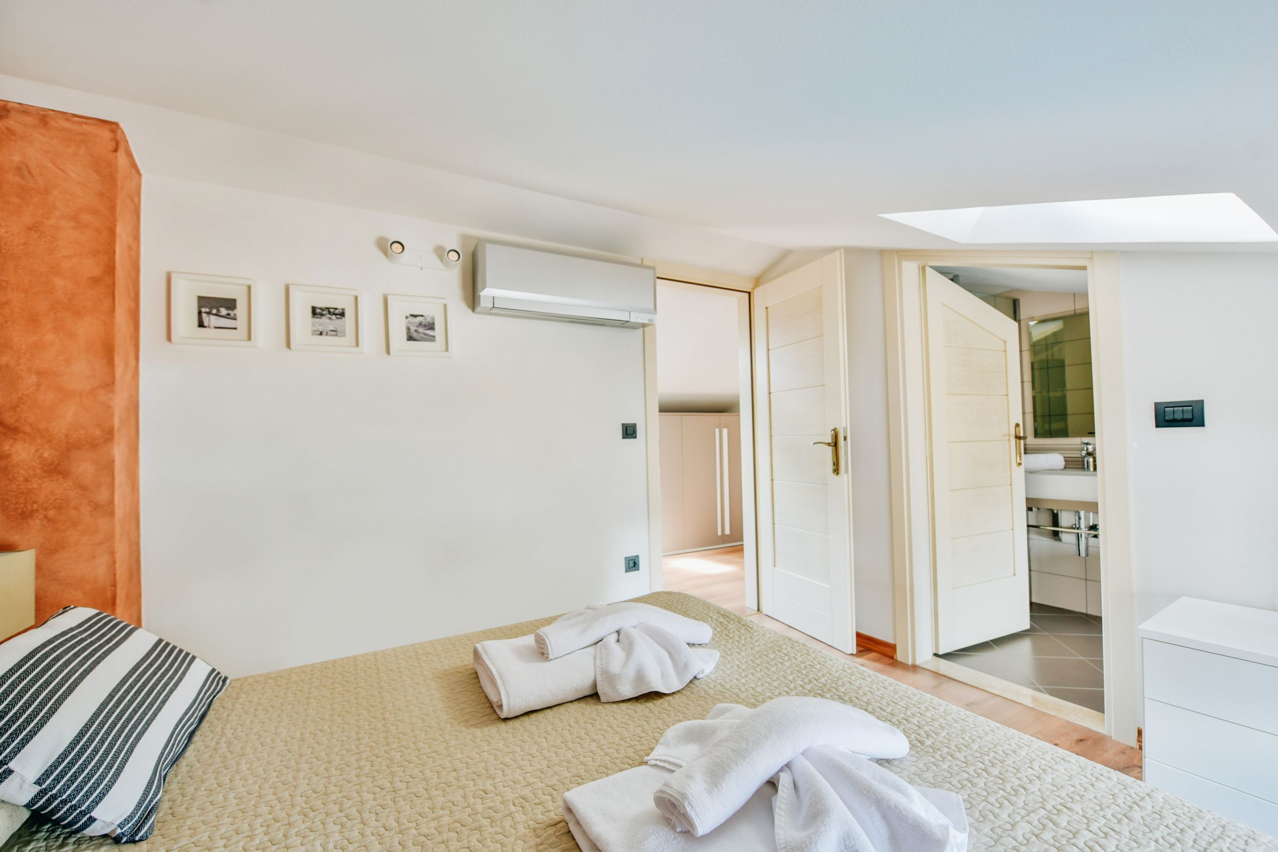 A bedroom with a bed, towels, a bedside table, a wall-mounted air conditioner, framed pictures, and an open door leading to a bathroom.