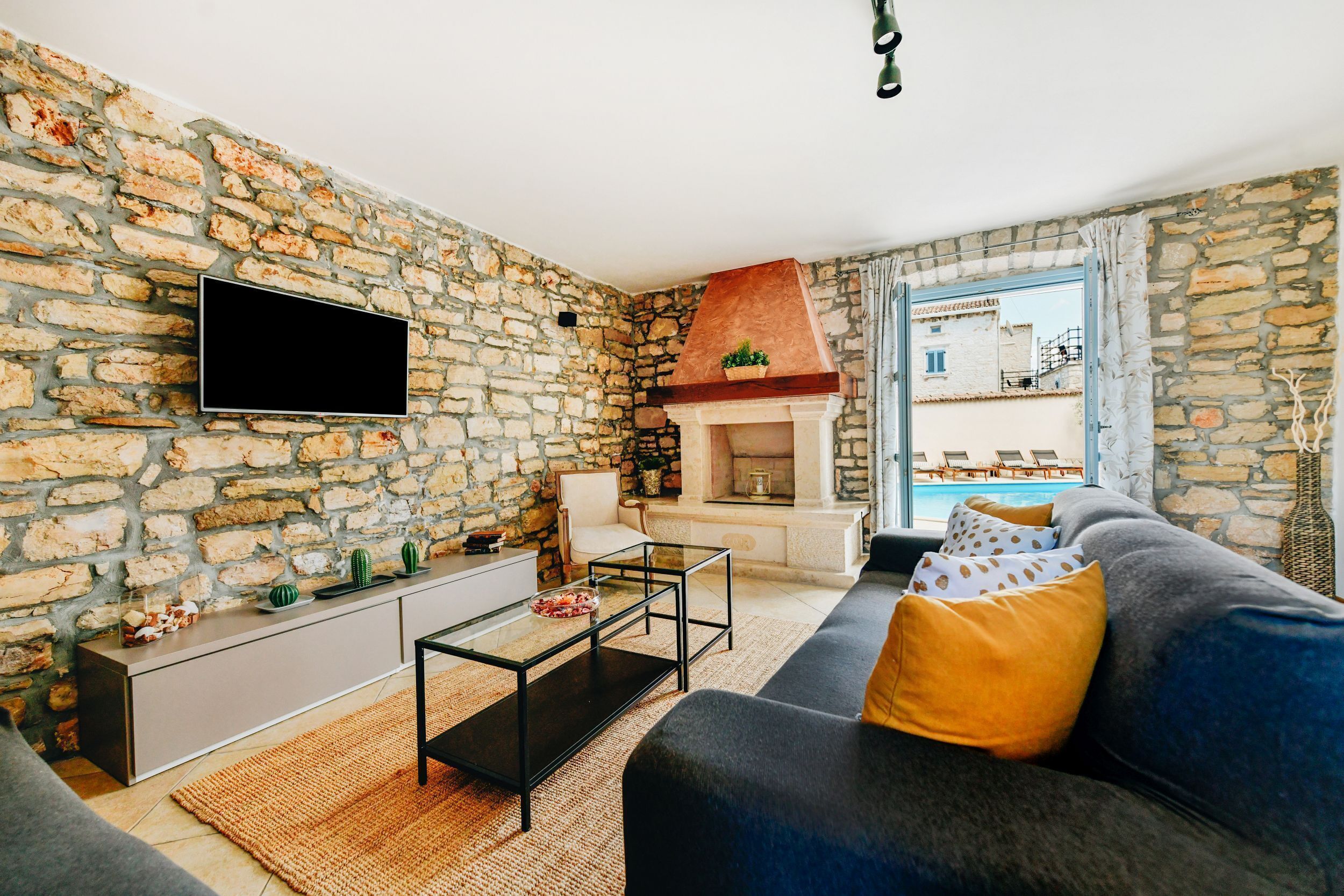 A living room with a stone wall, a flat-screen TV, a sofa with patterned cushions, a coffee table, and a fireplace.