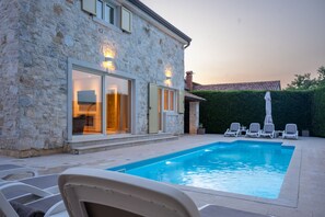 A stone building with a swimming pool, outdoor seating, and a hedge.