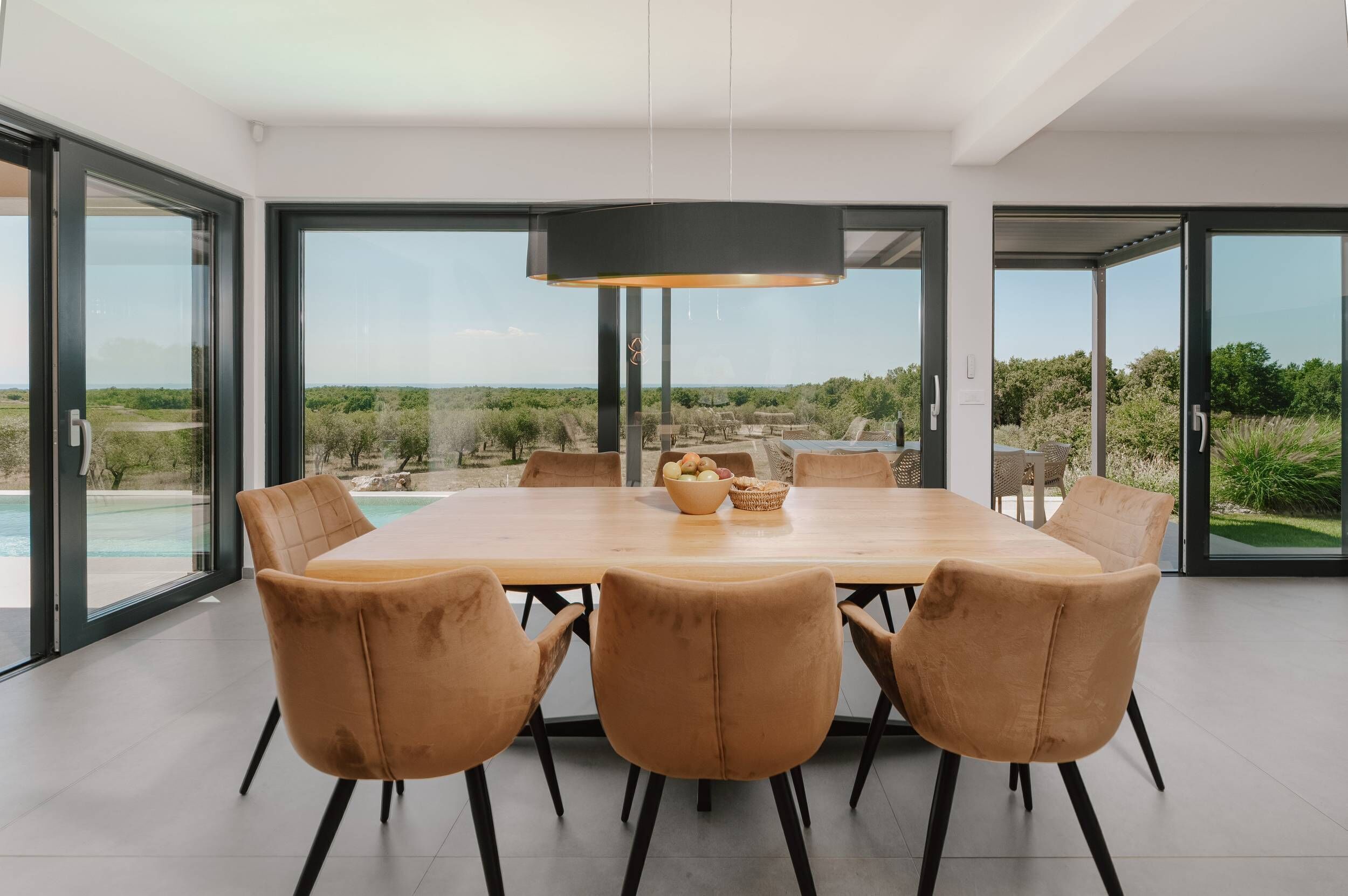 A modern dining area with a large table and comfortable chairs, large windows offering a view of the outdoors, and a hanging light fixture.