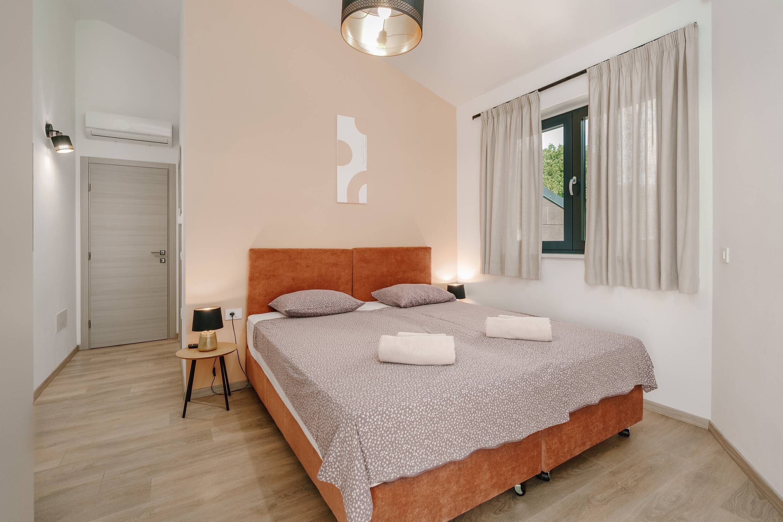 A bedroom with a bed, a bedside table, a lamp, a window with curtains, and a wall-mounted air conditioner.