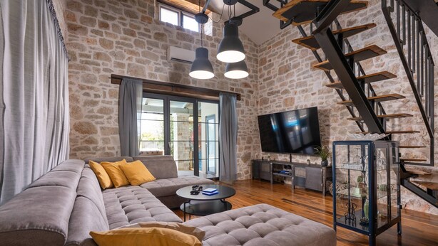 A modern living room with a stone wall, a large sectional sofa, a flat-screen TV, and a staircase.