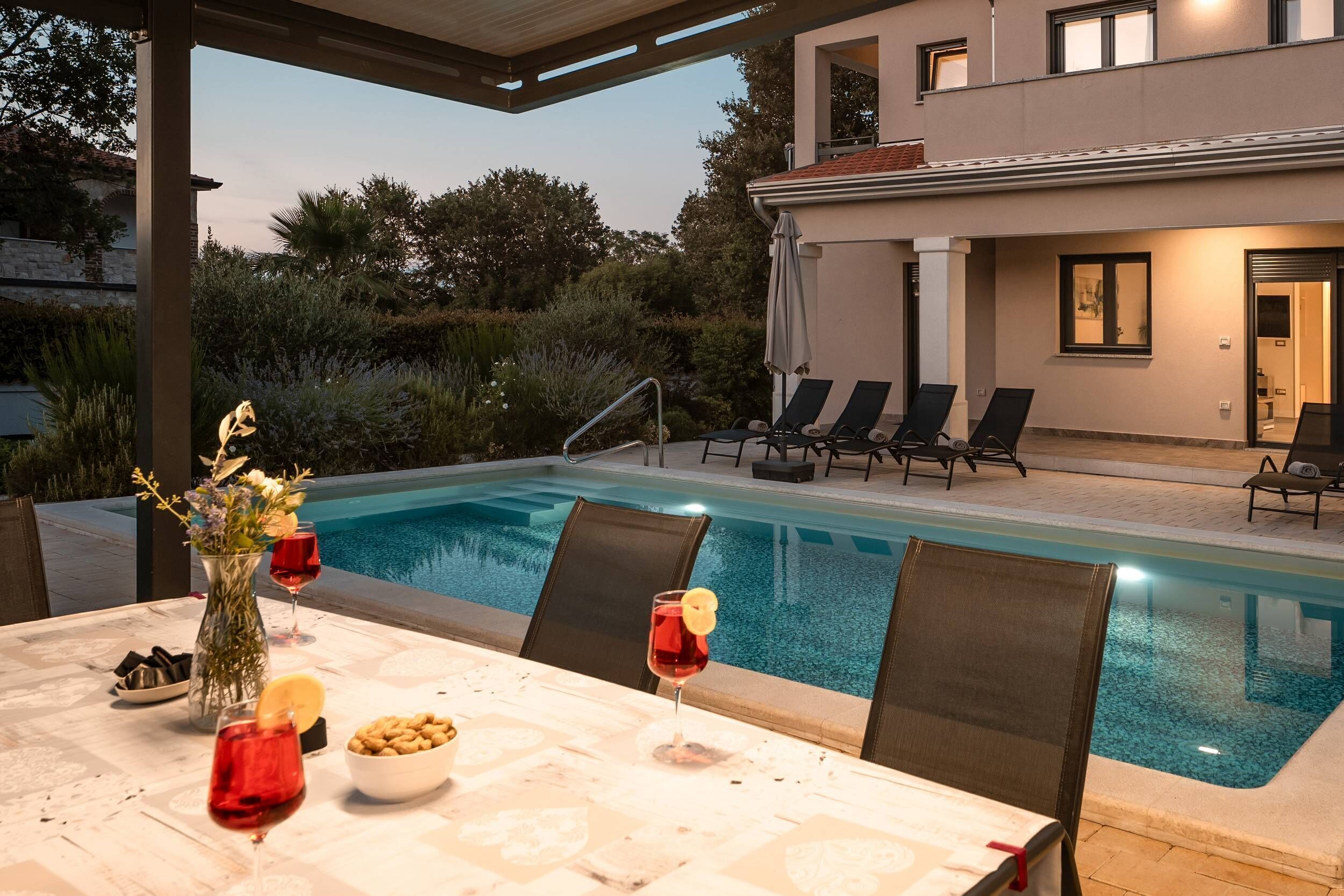 A poolside dining area with a table set for two, a vase with flowers, and a bowl of nuts.
