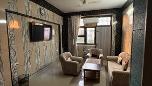 A spacious living room with a flat-screen TV mounted on the wall, a ceiling fan, and a large window with curtains. - Awaraj Home Stay (Agra)
