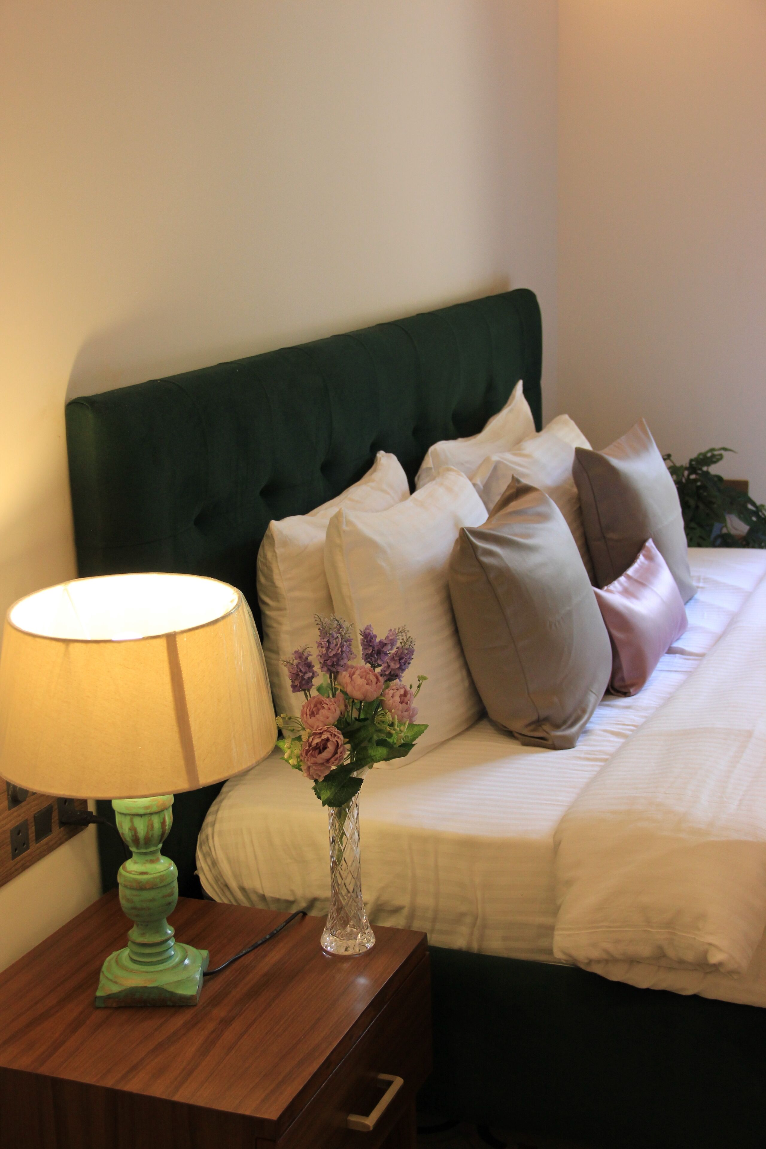 Una cama impecablemente arreglada con cabecera verde, varios almohadones, un florero con flores y una lámpara sobre una mesita de noche de madera.