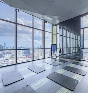 Amplia habitación con ventanas grandes que ofrecen vista a la ciudad, colchonetas de ejercicio en el suelo y un reloj digital mostrando la hora.