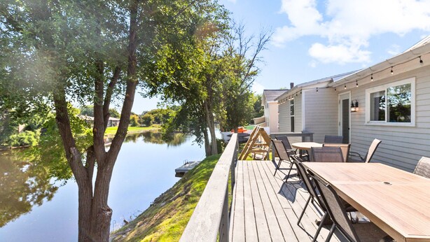 A wooden deck with outdoor seating overlooking a calm lake, connected to a building with large windows.