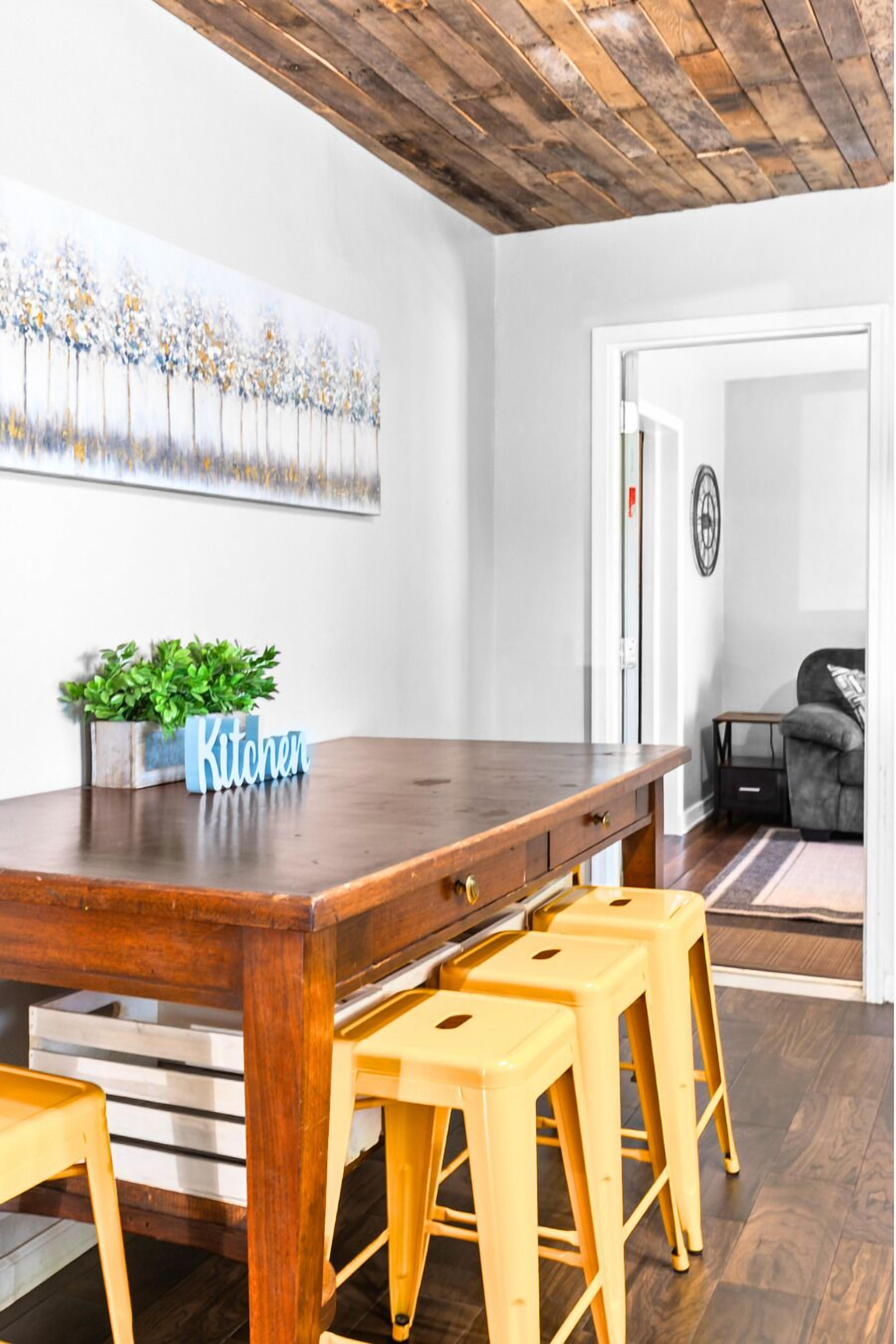 A wooden dining table with a 'Kitchen' sign, three yellow stools, a potted plant, and a painting on the wall.