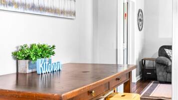 A wooden dining table with a 'Kitchen' sign, three yellow stools, a potted plant, and a painting on the wall.