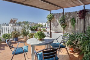 A terrace with a table and chairs, potted plants, and a view of buildings in the distance.