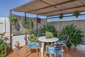 Een terras met een ronde tafel en stoelen, een houten schutting en hangende planten.