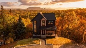 A two-story house with a dark roof, a front porch, and a staircase leading to the entrance. It is surrounded by dense trees with autumn foliage. - MountainSplash: Pool*MtnView*Pets*Games*HotTub*Wifi*FirePit (Sevierville)