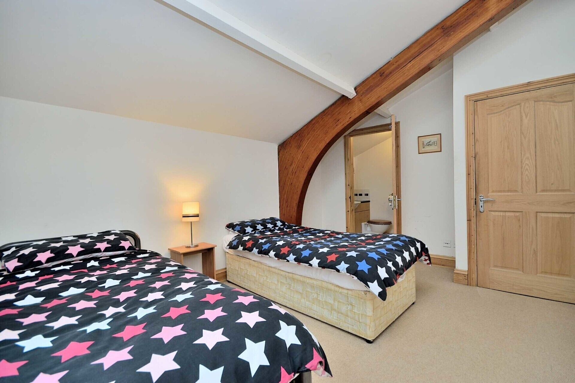 A bedroom with two beds, a wooden ceiling, a door, and a nightstand with a lamp.
