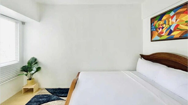 A bedroom with a bed, a window, a plant, a colorful painting, and a small wooden stool. - Cozy Staycation at Avida Iloilo (Iloilo)