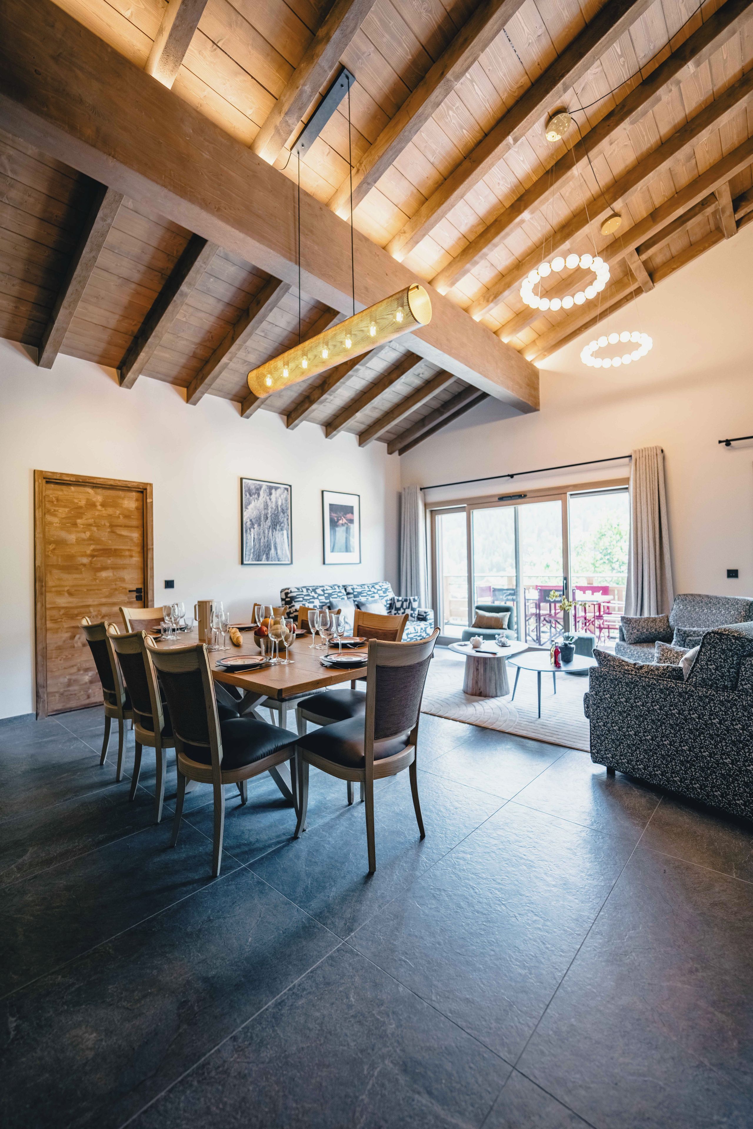 A spacious dining area with a wooden table set for a meal, flanked by chairs, in a room with a high ceiling and exposed beams.
